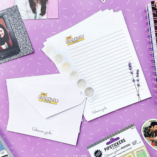 A styled flat-lay of Dragonfly Girls stationery on a purple background with white confetti. The set includes a lined letterhead pad, a matching envelope, a sticker strip with lavender illustrations, and a package of lip stickers.