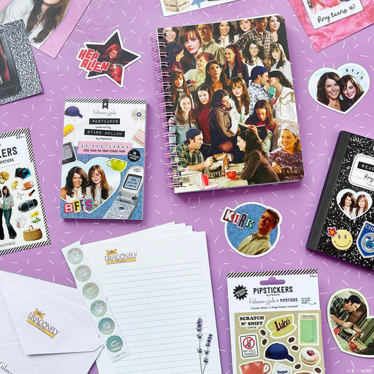 A styled flat-lay of Gilmore Girls stationery and stickers on a purple background. The set includes a spiral notebook with a collage of young women, a pack stickers, a notepad, and other matching stationery items.
