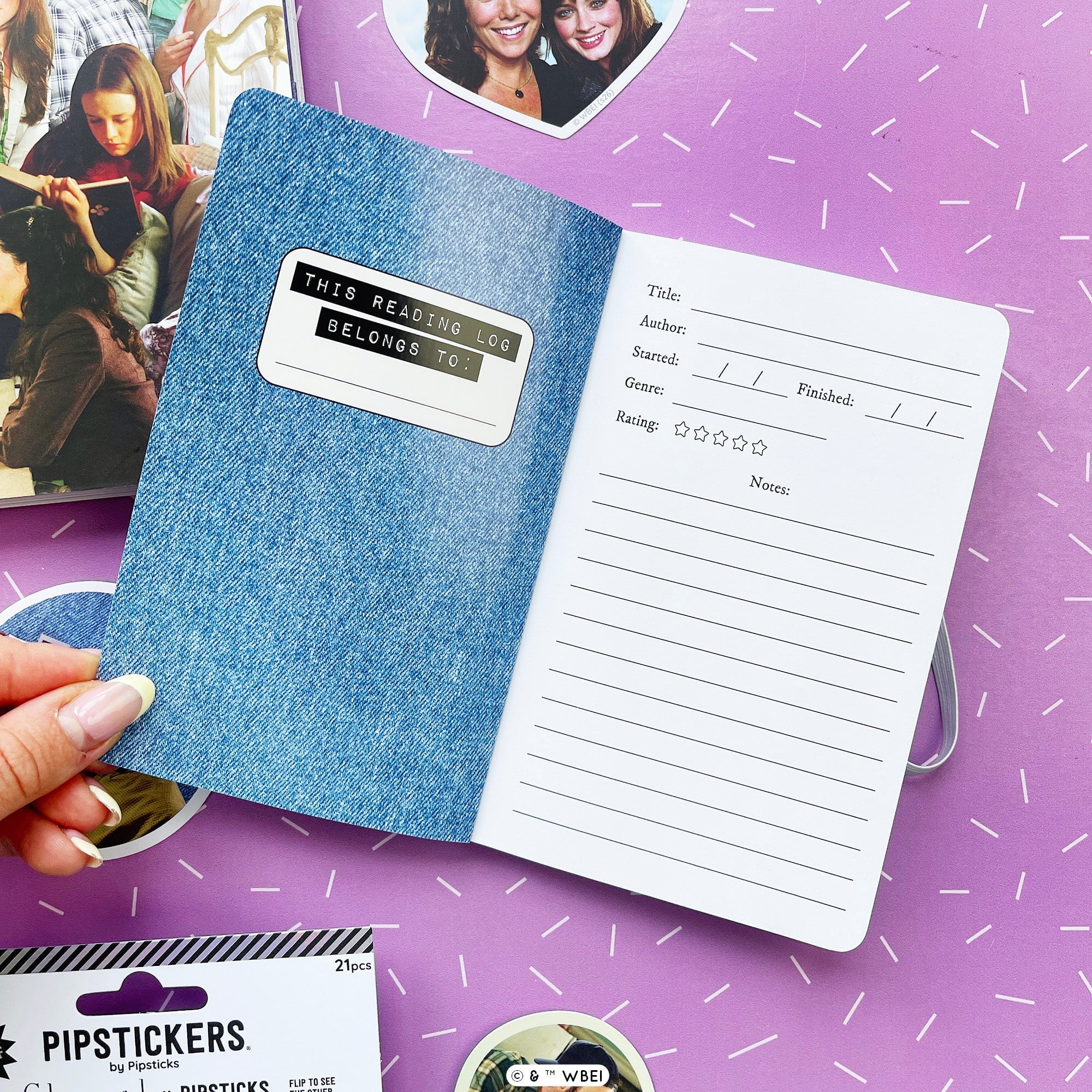 A hand holds an open mini journal with a blue denim-textured cover and white lined pages, placed on a purple surface with small decorative props around it.