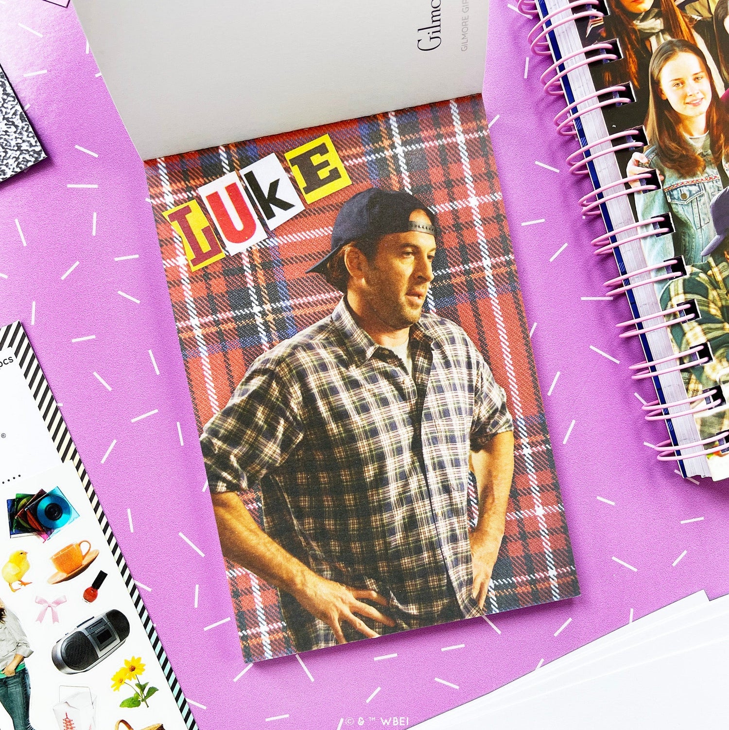 A styled flat-lay of a postcard in the postcard pack featuring a man in a plaid shirt and cap, placed on a purple surface with other stationery items around it. The card has a red, blue, and black plaid background with the word 'LUKE' in large colorful letters at the top.