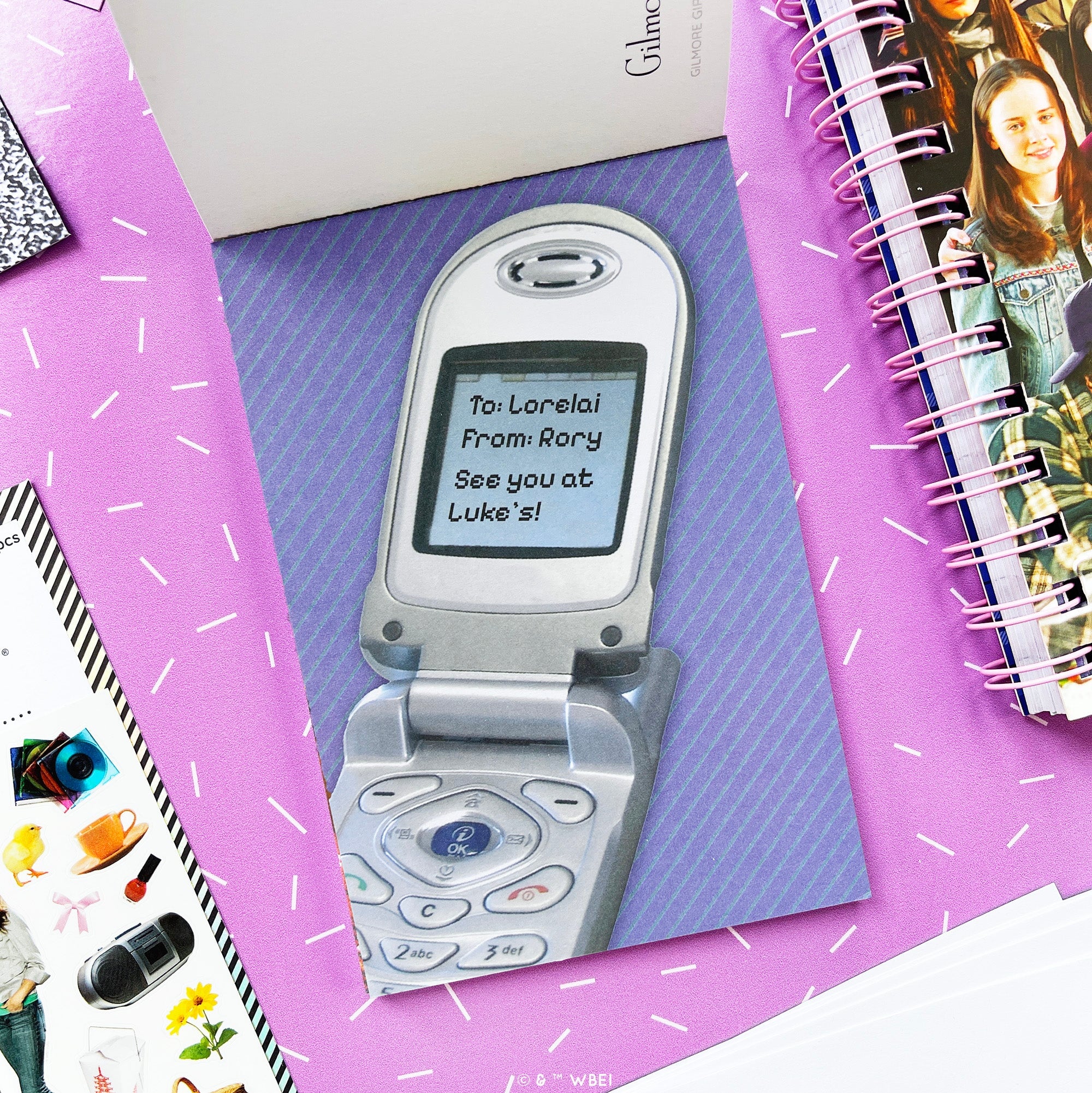 A personalized flip phone postcard featuring a printed image of a silver mobile phone with a message screen, displayed on a purple background with decorative stationery props.