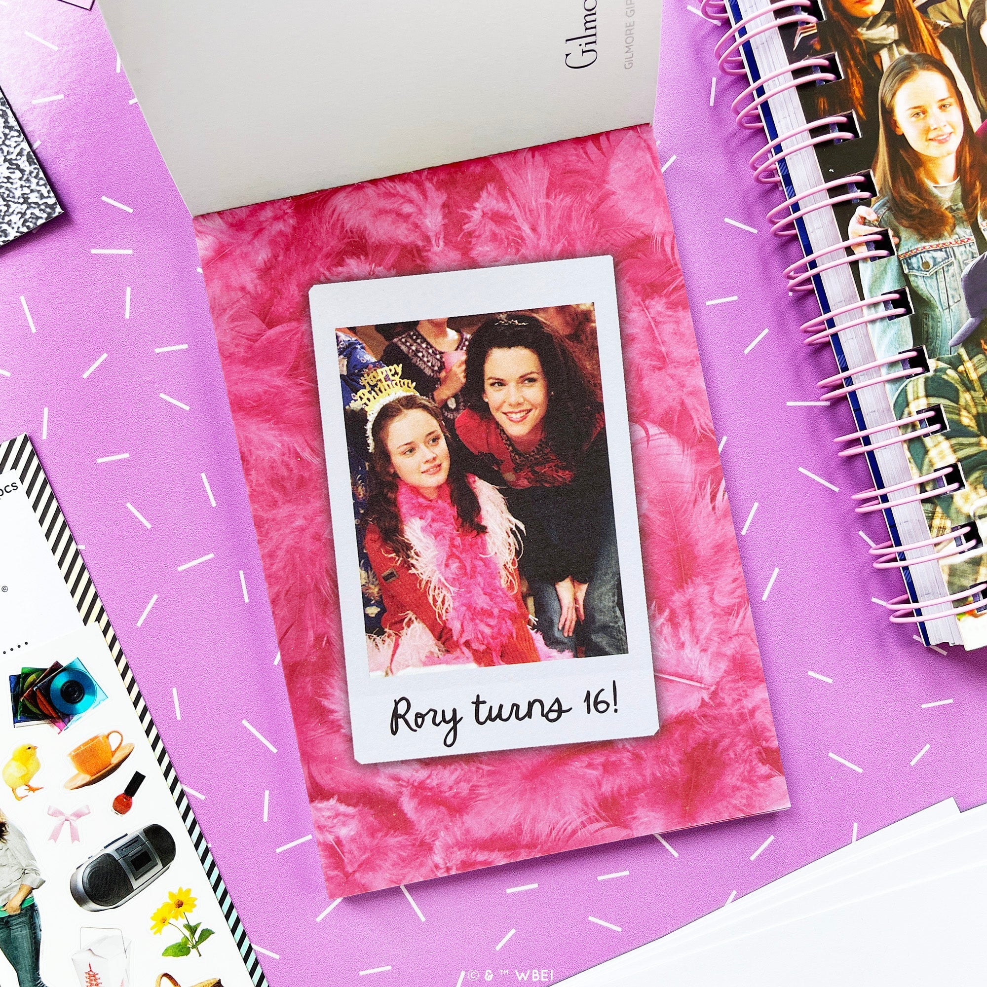 A styled flat-lay of a postcard in the postcard pack, feather-textured background and a central Polaroid-style photo of two girls with the text 'Rory turns 16!'. The page is surrounded by other stationery items on a purple surface.