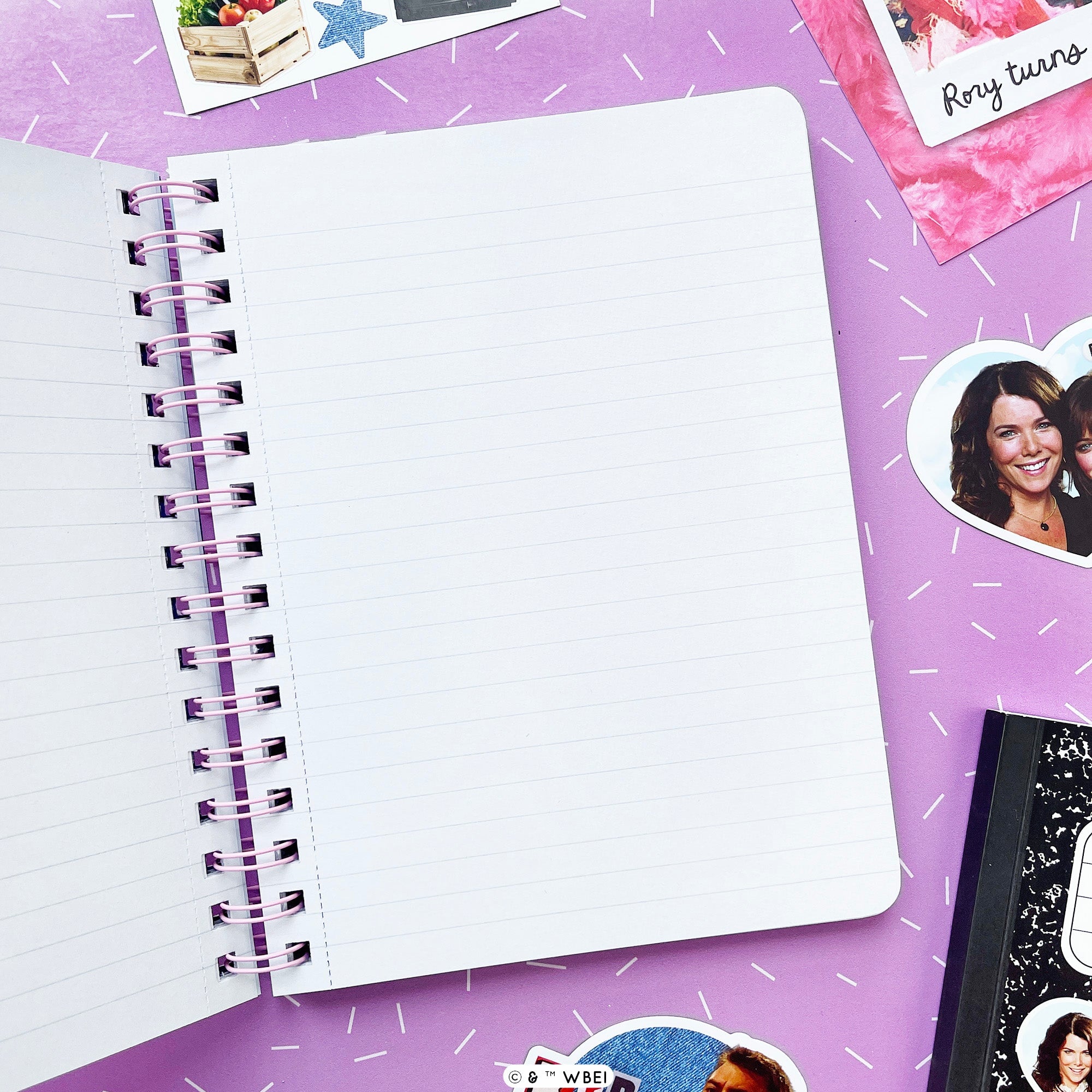 An open spiral-bound lined notebook with white pages and a purple binding, placed on a purple surface with small white dash marks. Other stationery items and photo stickers are partially visible around it.