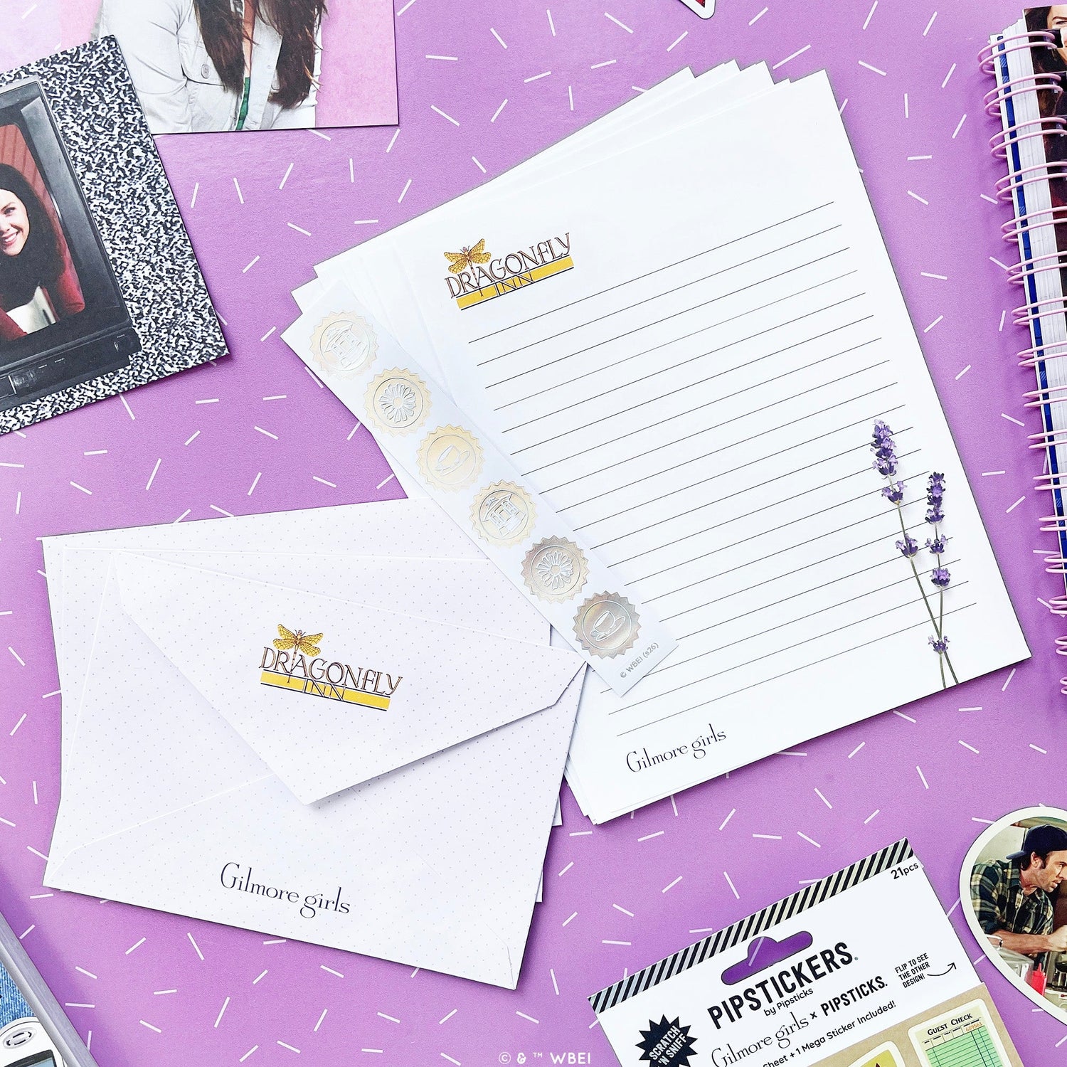 A styled flat-lay of Dragonfly Girls stationery on a purple background with white confetti. The set includes a lined letterhead pad, a matching envelope, a sticker strip with lavender illustrations, and a package of lip stickers.