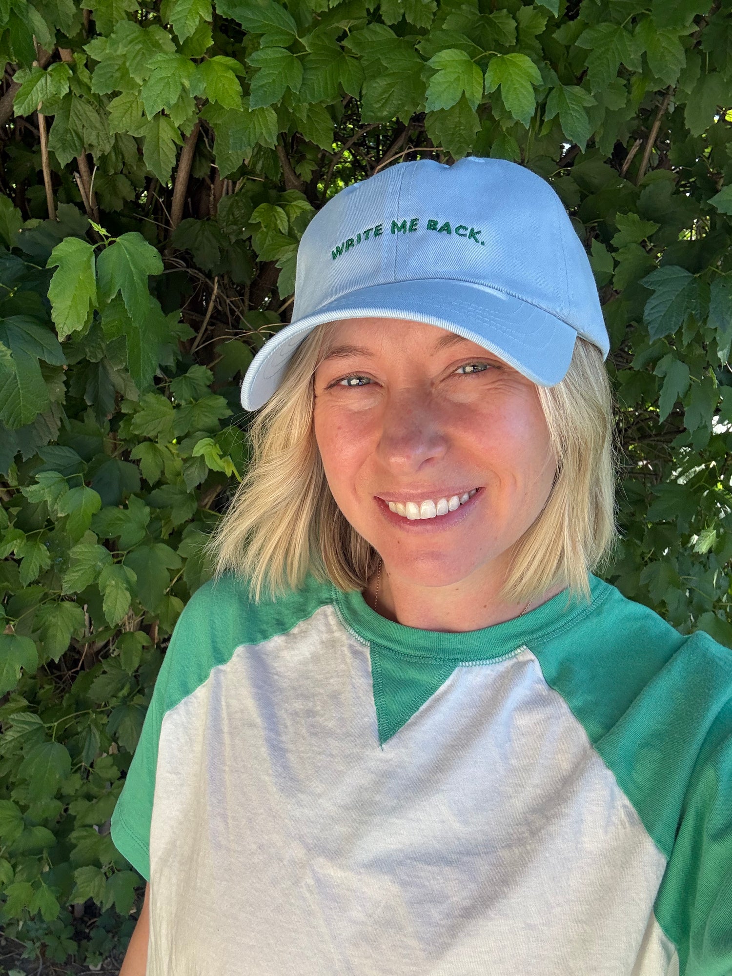 Person wearing a blue cap with 'Write Me Back' text, standing in front of green foliage.