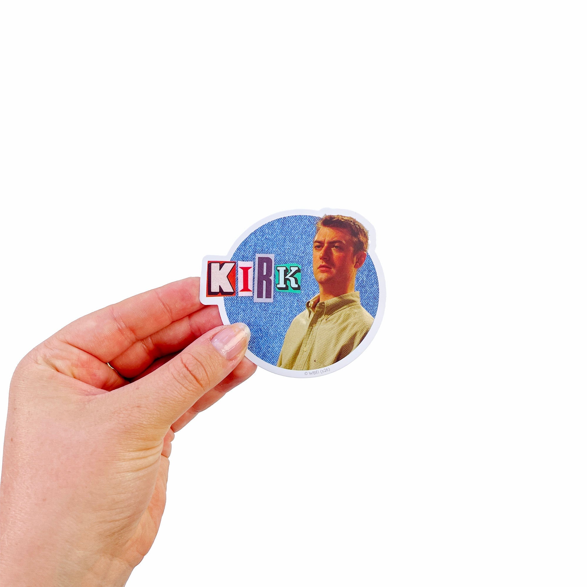 A hand holding a die-cut sticker featuring a portrait of Kirk from Gilmore Girls on a light blue textured background with the word 'KIRK' in colorful block letters.