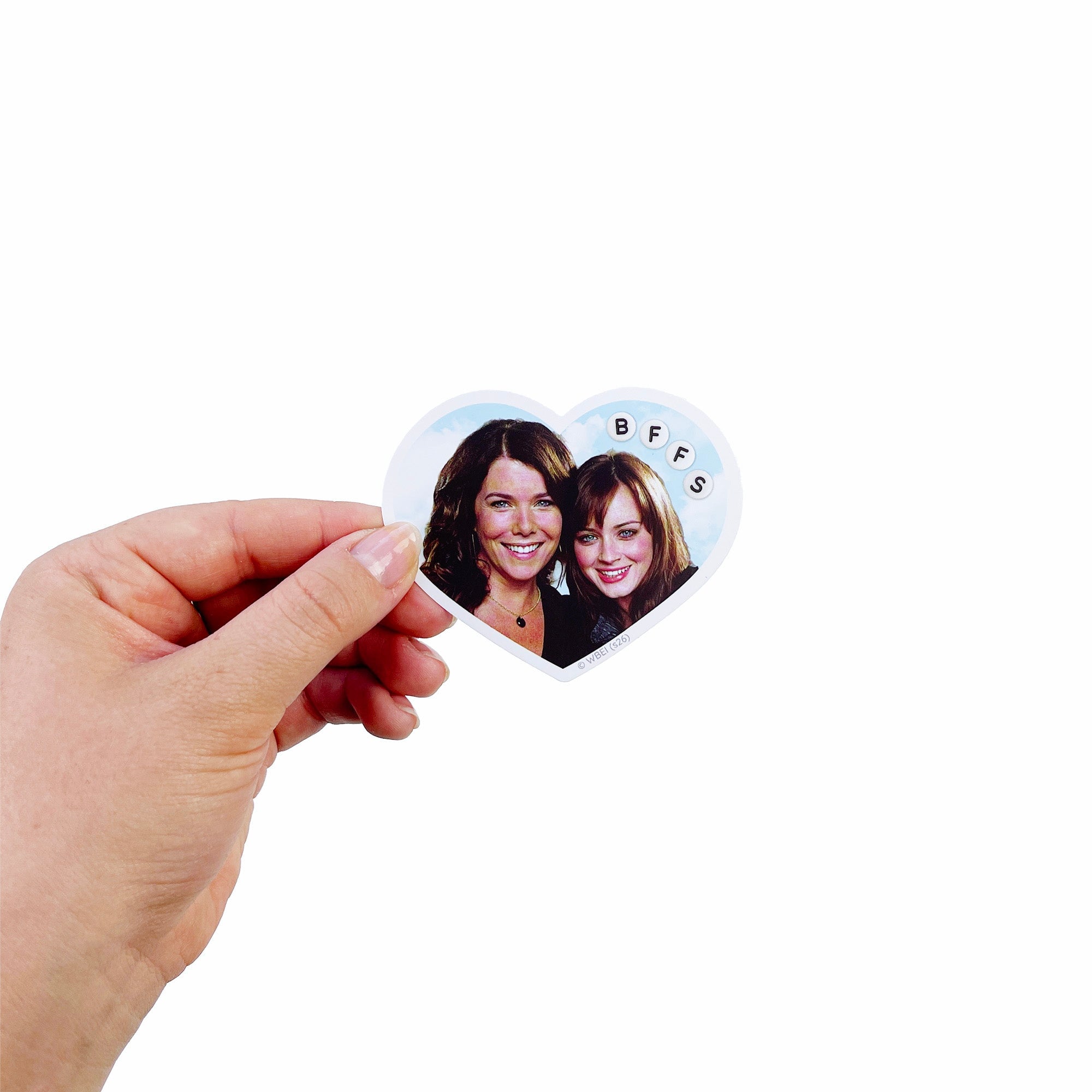 A hand holding a small heart-shaped sticker featuring a printed photo of two women with the letters 'BFFS' around the border, against a plain white background.